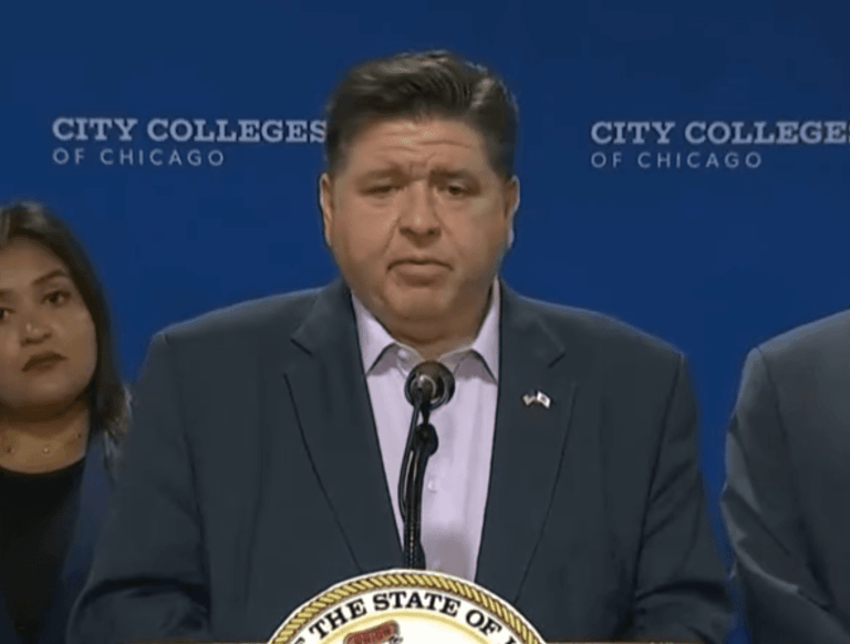 Gov. J.B. Pritzker stands at a podium during a press conference at the City Colleges of Chicago, addressing reporters with staff members behind him.