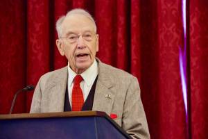 Senator Chuck Grassley speaking at a podium against red drapes during a press briefing on Capitol Hill, addressing concerns about the Department of Justice’s Arctic Frost investigation.