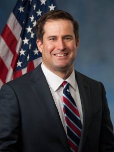 Official portrait of Rep. Seth Moulton (D-MA) in front of the American flag, wearing a dark suit, white shirt, and striped tie.