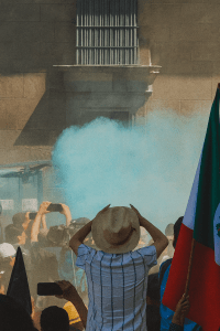 Protesters stand outside Mexico City’s presidential palace as blue tear gas rises, with a person in a straw hat facing the smoke and a Mexican flag visible on the right side.
