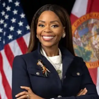 Smiling congresswoman standing with folded arms in a navy blazer in front of the United States flag and the Florida state flag