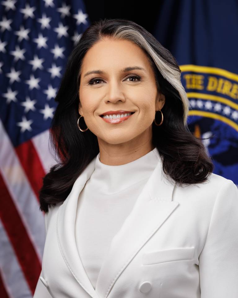 Official portrait of Director of National Intelligence Tulsi Gabbard wearing a white suit, standing in front of the American flag and the Office of the Director of National Intelligence emblem.