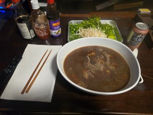 A large bowl of homemade beef pho with ramen noodles sliced beef rich dark broth and a platter of lime basil green onion cilantro and bean sprouts next to Sriracha Hoisin soy sauce and chop sticks on a napkin