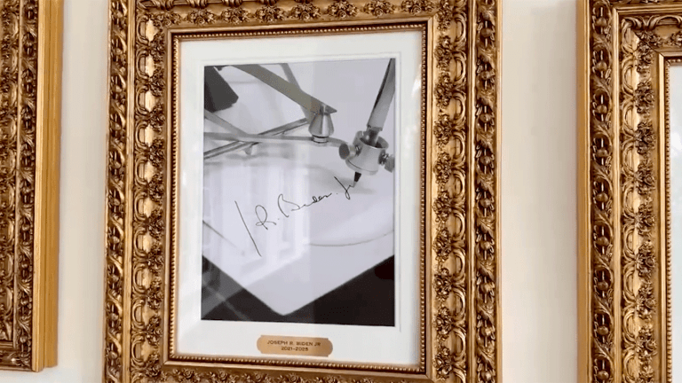 A framed photograph showing an autopen machine writing Joe Biden’s signature displayed in an ornate gold frame