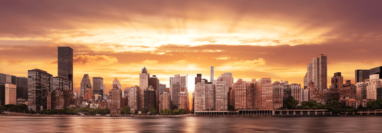 Panoramic view of the New York City skyline at sunset with orange and gold light over the East River.