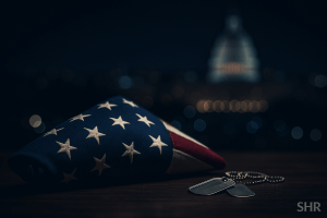 “Folded American flag with military dog tags in a dimly lit memorial setting, Washington DC skyline blurred in the background, tribute to fallen National Guard member Sarah Beckstrom.”