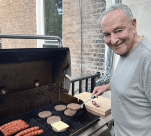 Chuck Schumer smiling while grilling hamburgers and sausages on a backyard grill