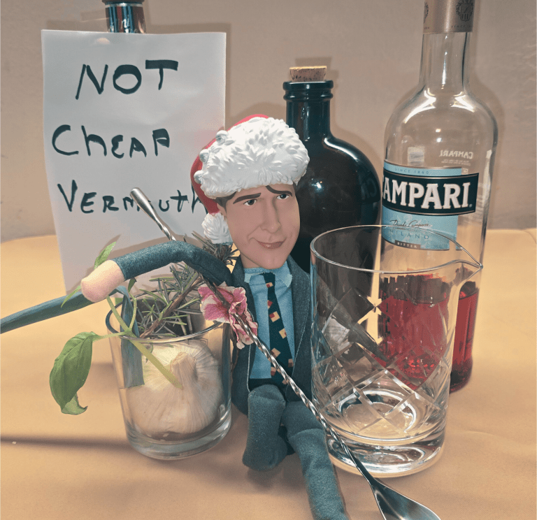 Boulevardier cocktail ingredients with Campari bottle, mixing glass, herbs, garlic and a holiday figurine beside a “Not Cheap Vermouth” sign