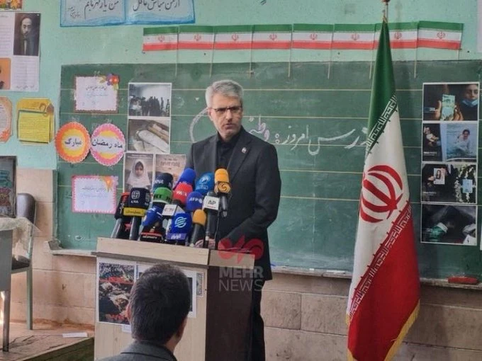 Iranian official speaking at a podium inside a classroom with Iranian flag and media microphones